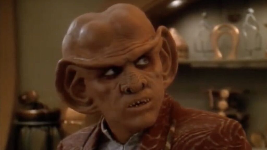 Quark the Ferengi bartender with glowing reflective eyes stands behind his bar counter in Star Trek: Deep Space Nine