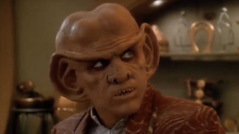 Quark the Ferengi bartender with glowing reflective eyes stands behind his bar counter in Star Trek: Deep Space Nine