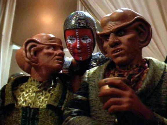 Three Ferengi characters with one wearing an ornate red and silver decorative mask in a Star Trek: Deep Space Nine scene