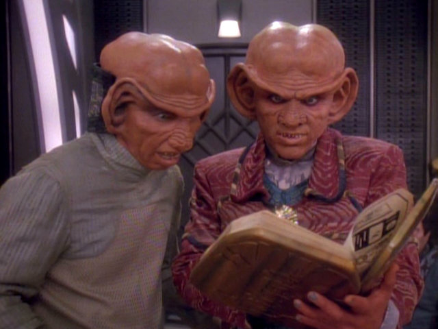 Quark and Nog lean over an open book together, studying what is likely the Ferengi Rules of Acquisition in Star Trek: DS9