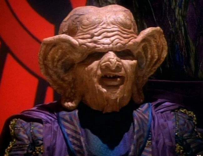 Close-up of an elderly Ferengi male with heavily ridged forehead wearing ornate purple and blue ceremonial robes in Star Trek: Deep Space Nine