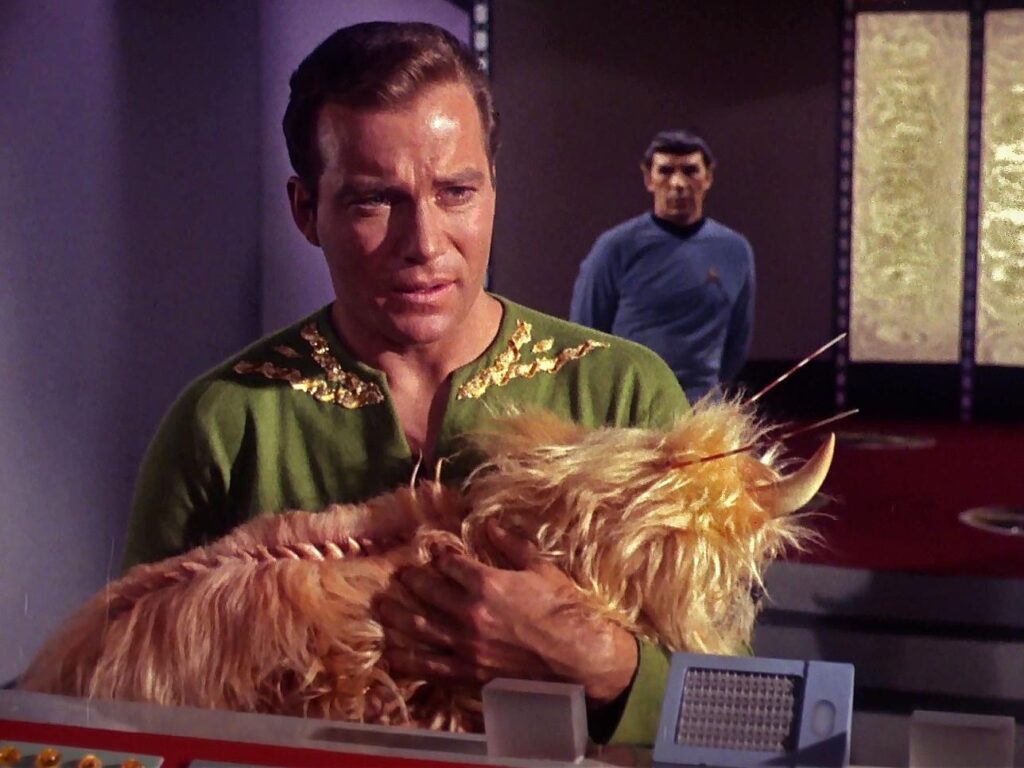 William Shatner as Captain Kirk in his green wrap tunic holding a Mugato (large fluffy alien creature with horns), with Leonard Nimoy as Spock visible in the background, from TOS episode 'A Private Little War'
