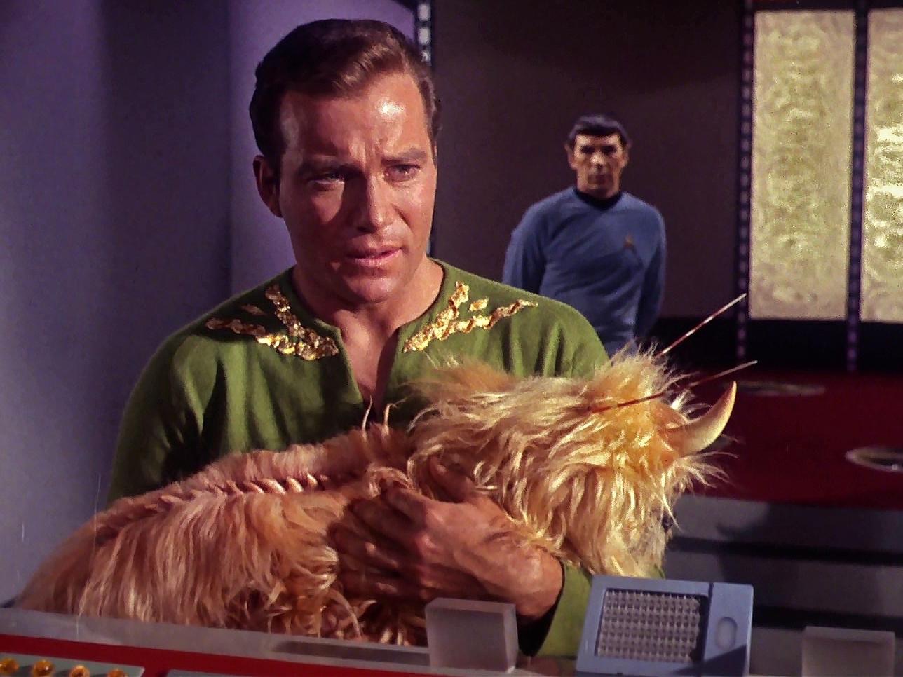 William Shatner as Captain Kirk in his green wrap tunic holding a Mugato (large fluffy alien creature with horns), with Leonard Nimoy as Spock visible in the background, from TOS episode 'A Private Little War'