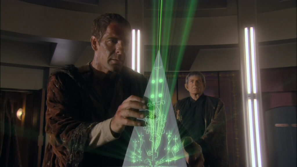A Starfleet officer studies a glowing green holographic display while two people watch.
