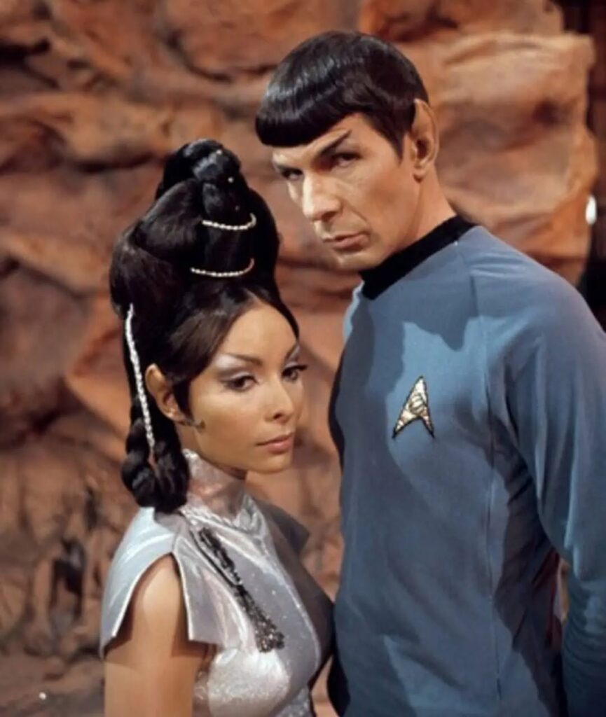 A Vulcan Starfleet officer stands closely beside a woman in white against a rocky background.