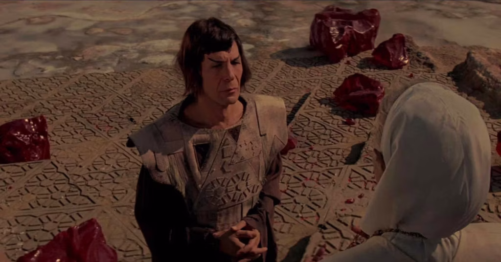 A Vulcan woman sits on patterned ground facing a robed elder during an outdoor ceremony.