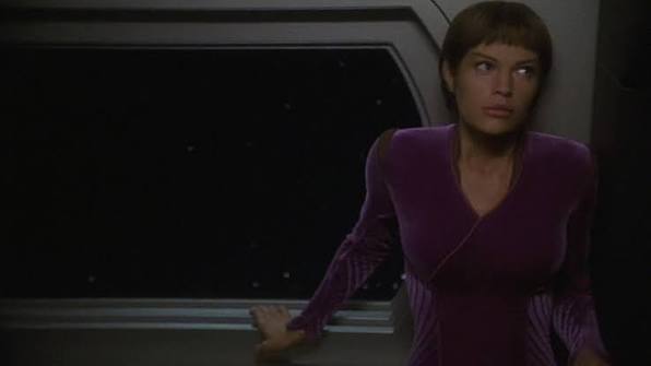 A Vulcan woman in a purple outfit stands beside a console on a starship bridge.