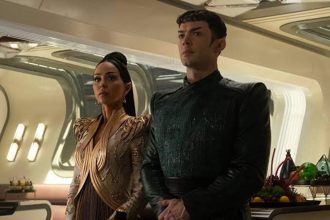 A formally dressed Vulcan couple stand together in a brightly lit room decorated for a gathering.