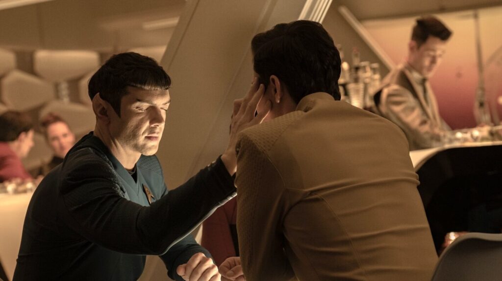 A Vulcan man reaches across a crowded table to touch another man’s face.
