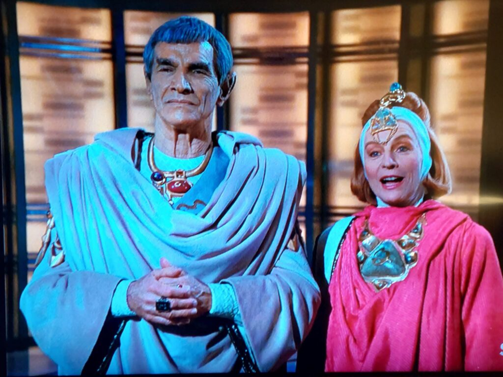 An elderly Vulcan man in blue ceremonial robes stands beside a smiling woman dressed in bright pink.