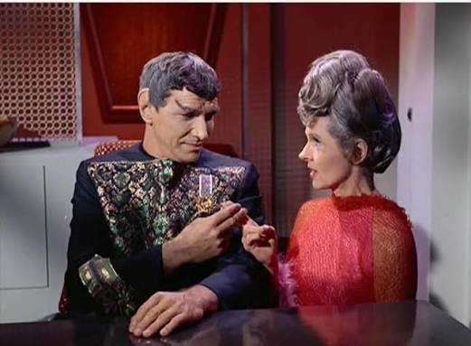 An elderly Vulcan man in ornate robes speaks with a woman wearing a red formal dress.