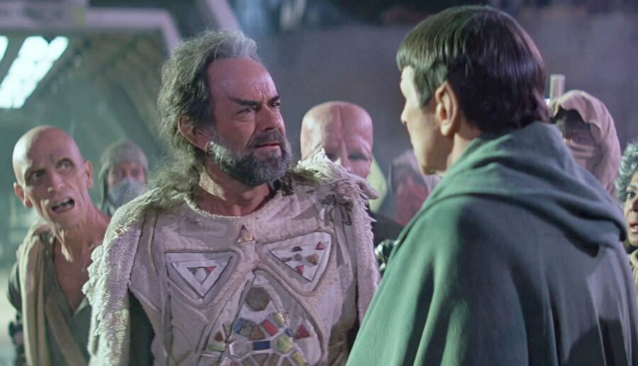 A bearded Vulcan elder in elaborate ceremonial clothing faces a younger Vulcan before a group of onlookers.
