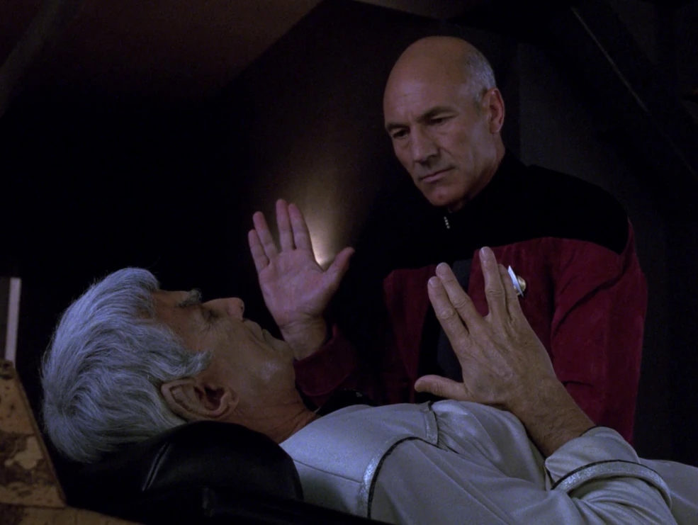 A bald Starfleet captain leans over an elderly Vulcan lying in bed during an intense telepathic exchange.