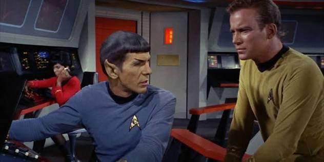 A Vulcan science officer sits at a bridge console while a Starfleet captain speaks to him.