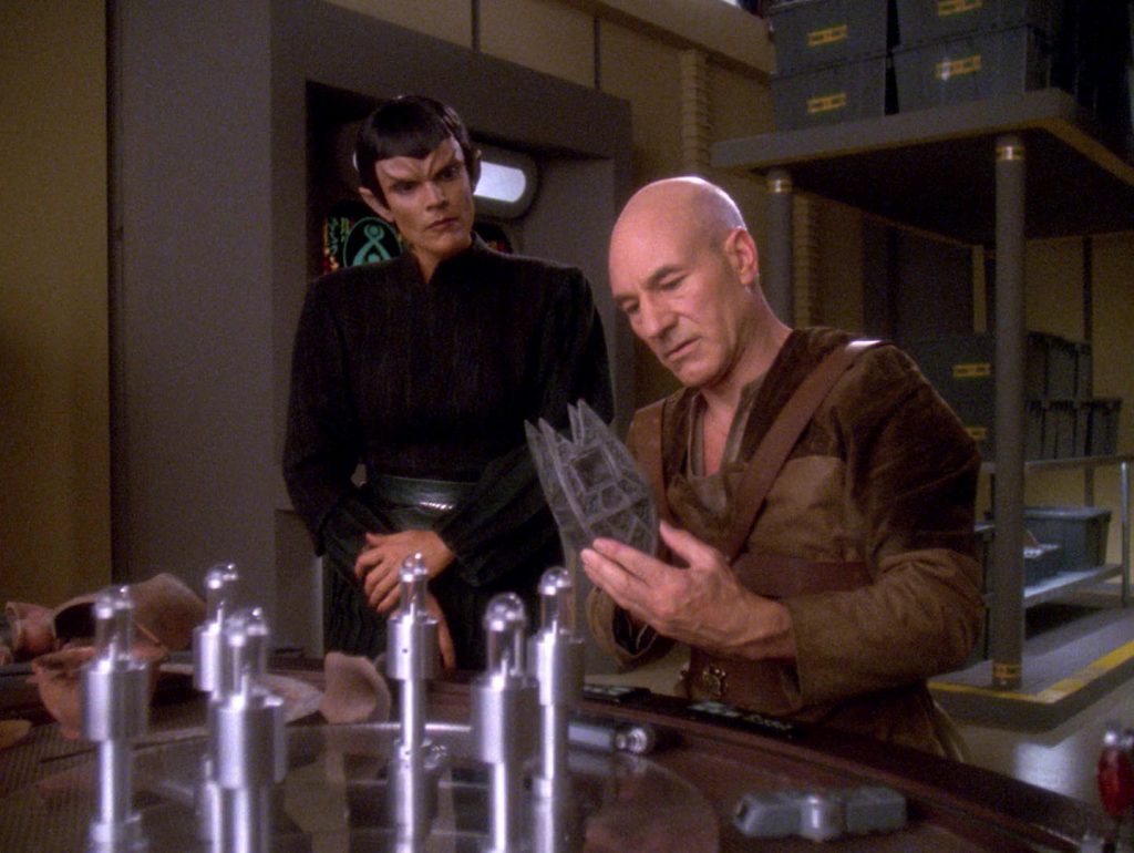 A Vulcan woman stands beside a seated man examining objects arranged on a table.