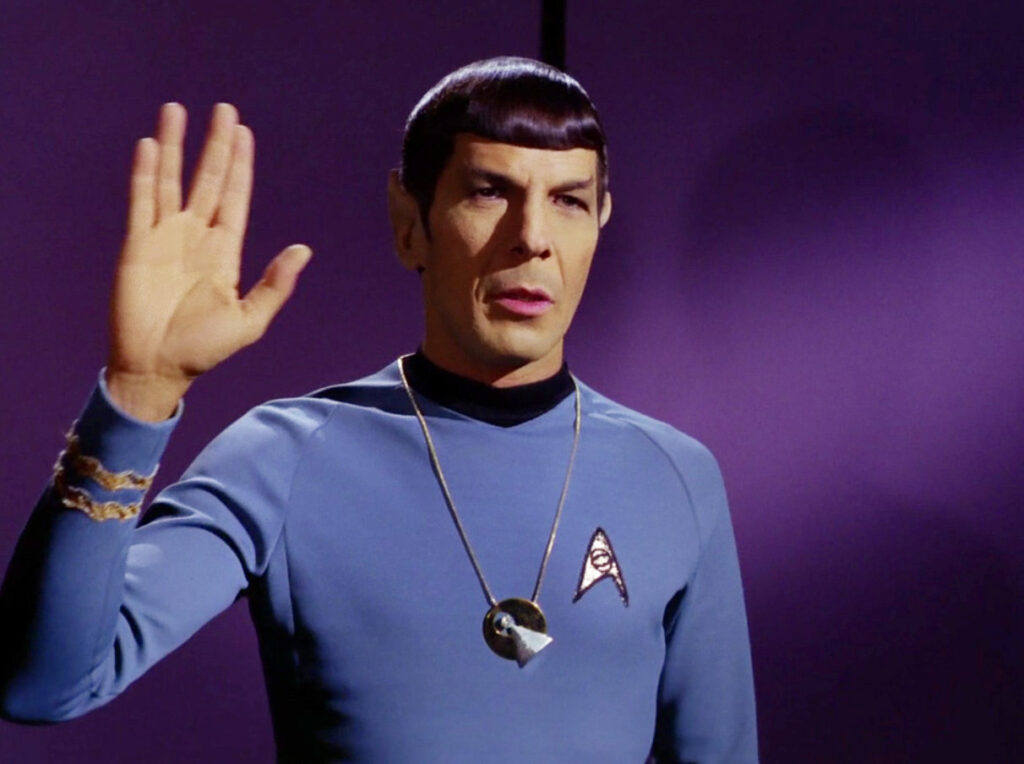 A Vulcan Starfleet officer in a blue uniform gives the traditional Vulcan salute.