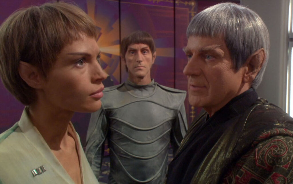 A young Vulcan woman speaks closely with an older Vulcan man while a guard stands behind them.